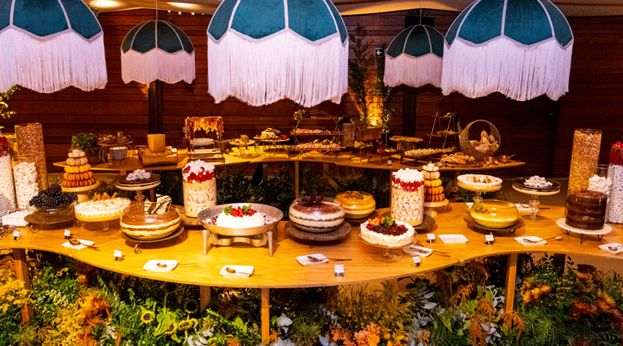 Ilha de sobremesas de casamento montada no Lajedo, com mesa farta de doces sob luminárias de franja